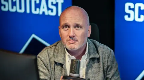 BBC Martin Geissler sits at a microphone in a radio studio, with scotcast logo in background, wearing grey shirt with grey stubble on face.