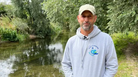 A man wearing a grey hoodie and baseball cap smiles as he stands on a grassy bank in front of a clear chalk stream with green trees behind him.