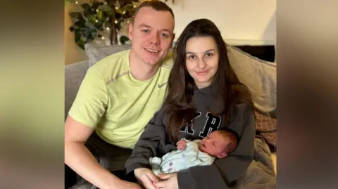 Alaw Llyfni Gwion, Alaw and Nanw sat together on a grey sofa. Gwion, on the left, wears a yellow Nike t-shirt. Alaw, on the right, wears a grey jumper and has long brown hair. She holds baby Nanw, who is wearing a blue baby grow. 