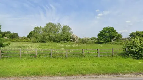 Google A large patch of land beside the A363. The picture is taken on a sunny day in summer, and the trees and grass is bright green and overgrown. There is a wooden fence blocking off the site from the road.