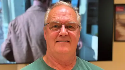 Steve Barrett smiling for the camera. A screen is showing some old film in the background.
