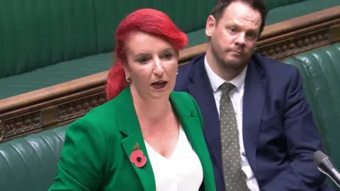 Parliament Live Transport Secretary Louise Haigh speaking at the Houses of Parliament. She is standing up. She is wearing a white top and a green blazer. A man in a dark blue suit is sitting behind her.