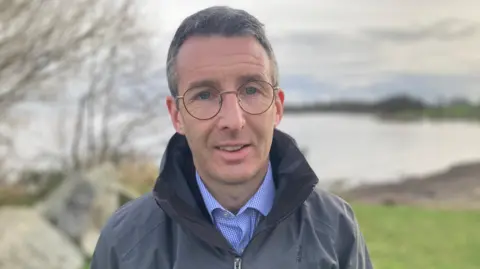 Andrew Muir standing on the shore of Lough Neagh in February 2024.  He has short, dark, greying hair and glasses.  He is wearing a waterproof navy coat with the collar turned up, over a blue shirt.