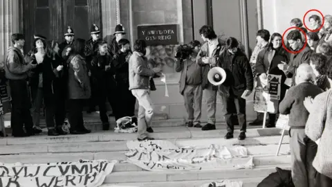Chris Reynolds This is a wide, black and white photograph of the Welsh Office during the 1991 protest. A number of people are gathered in front of the main door. There are police officers guarding the door. There is graffiti on the wall. Rhun ap Iorwerth can be seen to the right of the photograph.