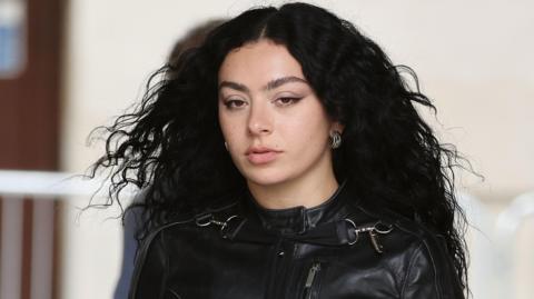 Charli XCX in London in June 2024. Charli is a woman in her early 30s with long curly black hair worn loose. She has brown eyes and wears a leather biker-style jacket. 