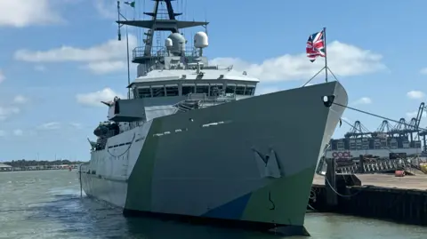 The large grey ship docked in Harwich. It has a Union Jack flag flying at the front.