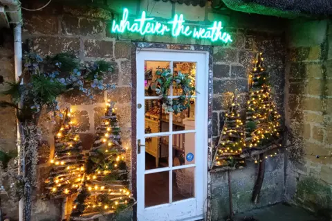 Ian W / BBC Weather Watchers Christmas lights around a door in Broadway, Worcestershire. A lit sign above the door reads: "Whatever the Weather"