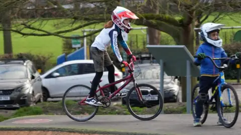 Child on a bike in a red helmet with red gloves and another child in a white helmet wearing blue. The background is a park like enviroment.
