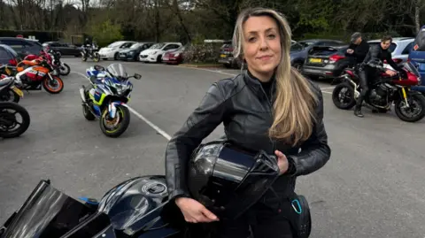 Ed Cook/BBC Stass Daniells pictured with her bike outside a café in Dorking. Stass is wearing all black and is holding her helmet. She has long brown/blonde hair and is stood beside her motorbike. A number of motorbikes can be seen in the background.