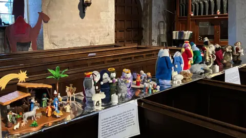 A row of four nativity scenes inside the church, amongst the pews. Two are knitted figures, another is in fabric, and another is Playmobil.