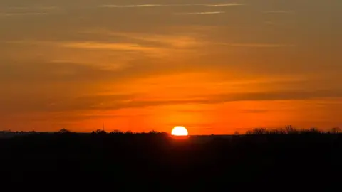 FLOWER GIRL/WEATHERWATCHERS The sky turns orange as a sun beings its rise above Royston, in Hertfordshire. Half of the image - the part of the image below the sky - is black.