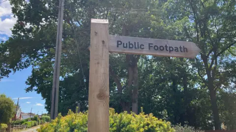 BBC/Emily Coady-Stemp A wooden public footpath signpost in the Surrey village of Normandy, framed against several trees and a bush.