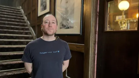 BBC Julio Romero Johnson, in a black t-shirt, is standing at the bottom of a carpeted staircase with photos on the wall next to it. He has long straight brown hair tied at the back, blue eyes and small silver hoop earrings. 