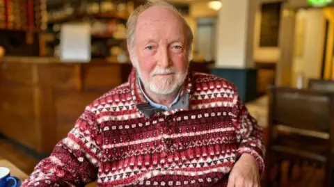 Martin Giles/BBC Dave McAllister is sitting in a pub with the bar in the background. It is a wood paneled bar. Dave is wearing a red and white patterned fleece, with a blue shirt underneath. He has a beard and is smiling at the camera. 