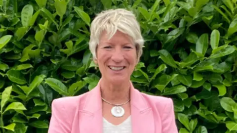 Woman with short blonde hair wearing a pink jacket and white top with a green bush in the background