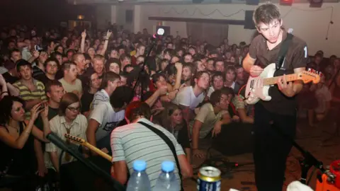 Getty Images Alex Turner, Arctic Monkeys frontman, pictured playing a guitar on stage. He has brown hair and is wearing a brown polo shirt.