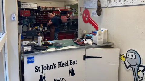 A man works in the back of a workshop, he is making repairs to shoes stood in front of machinery. There is a grey key cutting sign on a white wall and a trade counter with a number if items on top of it, including a shoe, card machine and an aerosol. 