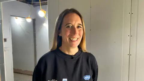 Photo of guest Rachel Jones, CEO of Bath Cats and Dogs Home. She is standing in the corridor of a refurbished building which is still under construction. She is wearing a blue t-shirt and has mid-length blonde hair 