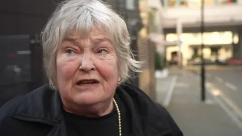 A woman in a black coat with short grey hair, looks up at the building from outside 