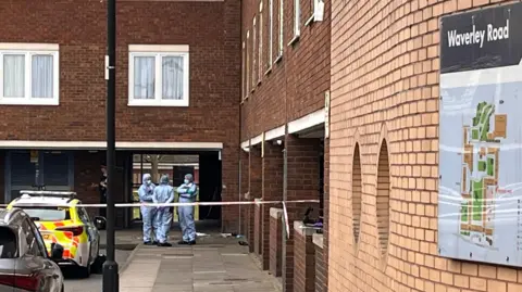 BBC Three forensic officers standing behind police tape wearing blue forensic overalls. There is a sign on a brick wall of a building saying Waverley Road