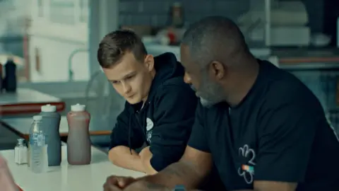 Jayden and Idris Elba sit next to each other at a table in a cafe where they talk about how Jayden has benefitted from the Cirv scheme. Jayden is wearing a black hoodie with his sleeves rolled up while Idris wears a black T-shirt.