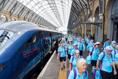 PA Media Dozens of people wearing bright blue polo shirts and purple backpacks walk down the platform of a railway station beside a blue train which has a name badge reading: "Jean Bishop (The Bee Lady)". The station has yellow-brick, arched walls and a huge arched glass roof. A station clock can be seen above a girder to the right.