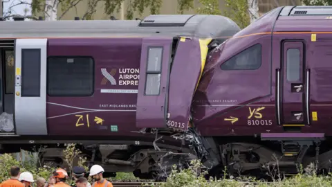 Two damaged trains, the front of one purple train is crushed into the end of another, with severe impact damage visible around the cab area.