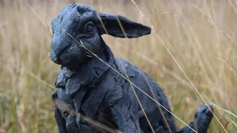 A mostly realistically-shaped hare sculpture in black with ears back in long grass