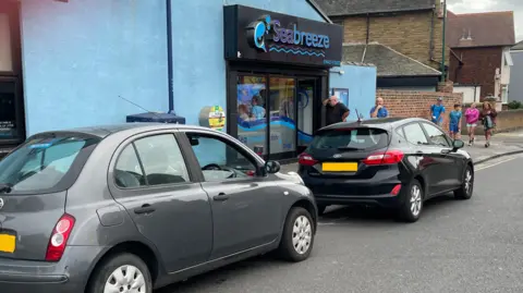 Two cars - one grey and one black - parked outside a fish and chip shop.