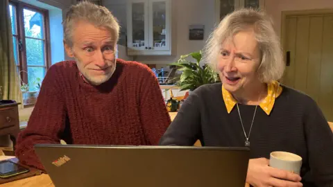 A man and woman, both with greying hair, are sitting at the kitchen table of their home, looking in frustration at a laptop. He is wearing a dark red jumper, she a navy blue one over a yellow blouse. 