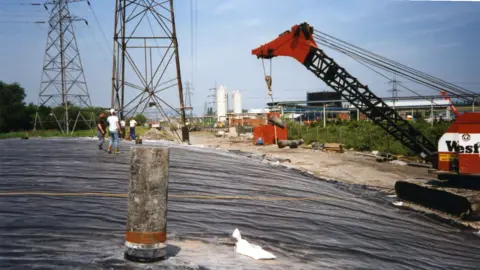Stephen Constance A work site showing a black plastic sheet covering over a mound of something. A crane is on the right and people are stood on top of the sheet in normal clothes, they do not appear to be wearing PPE