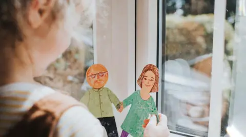 An over the shoulder shot of a young girl holding a cut out paper drawing of what appears to be a mother and father holding hands. She is holding the drawings looking out of a window into what appears to be a garden. 