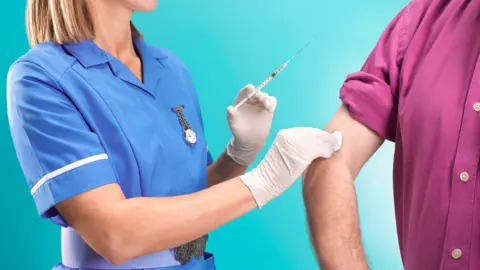 Female nurse giving flu jab to senior man. The nurse has short blonde hair wearing a blue nurse's uniform and rubber gloves, holding a syringe. She is pressing a cotton ball on the man's arm. He is wearing a pink shirt.