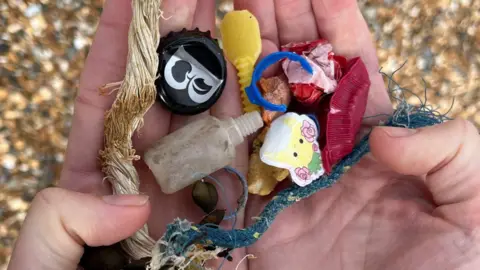 Various pieces of plastic and other beach waste in two open palms of a person's hand. Beneath the hands are pebbles on Brighton's beach