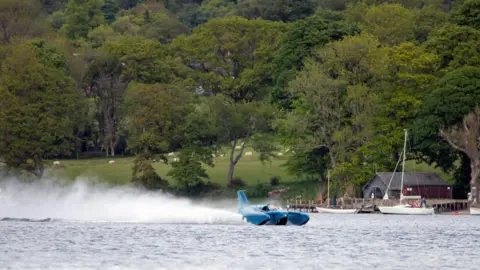 Bluebird K7 "planes" on Coniston Water. The blue craft is throwing up a large spray behind it as it glides on top of the lake's surface. On the far shore, sheep are grazing on an incline.