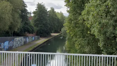 A Google Maps image of a canal. We can see a metal gate at the bottom of the screen. Then the canal bends off to the right with some birds swimming in it. There is a wall along the canal with graffiti on it. To the right of the screen there are trees and bushes. 