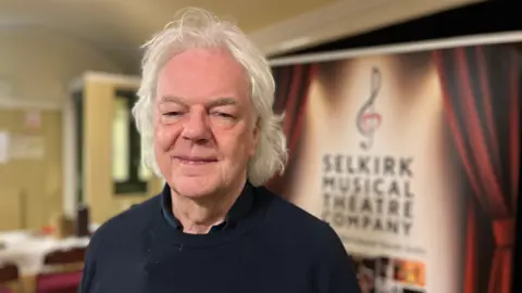 A man with grey, shoulder-length hair, wearing a black top, looks at the camera.