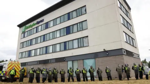 REUTERS/Hollie Adams A row of police officers with riot shields and wearing helmets stand in front of a hotel building. To one side is a police van.