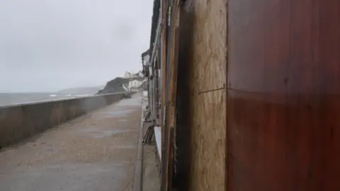The picture shows a seafront walkway on a grey, rainy day. On the left side, waves are rolling in towards a long concrete sea wall that runs the length of the image. Beyond the wall, the coastline curves towards a cluster of buildings in the distance, including one on a small rise overlooking the water.