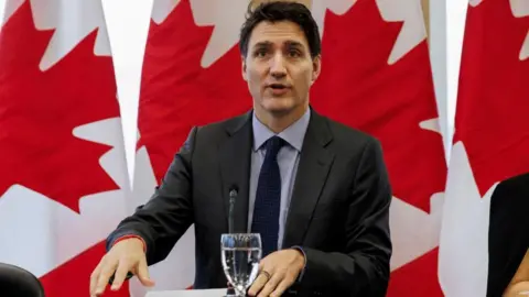 Reuters Justin Trudeau speaks in front of a microphone with Canadian flags in the background.