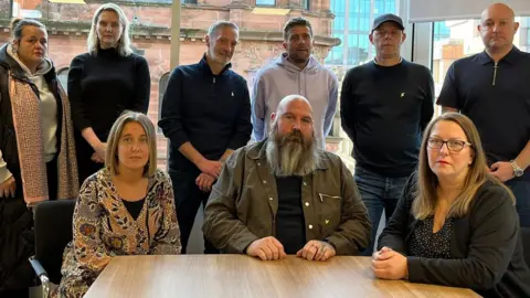 Seven family members gathered in a room with two lawyers. Seated at a table are, from left to right, Louise Slorance, Mark Bisset and Maureen Dynes. Standing behind them are, from left to right, Samantha McDowall; lawyers Stephanie Spencer and Patrick McGuire; Kevin McDowall; Kenneth Murdoch; and David Campbell.