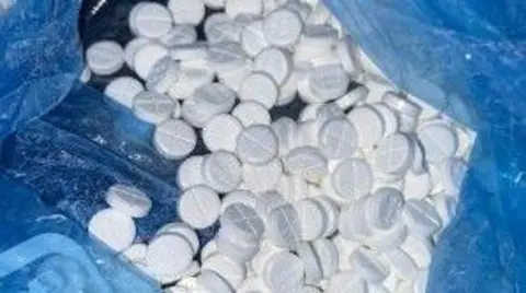 National Crime Agency Close up of small white round pills with crosses marked on them, in blue plastic.