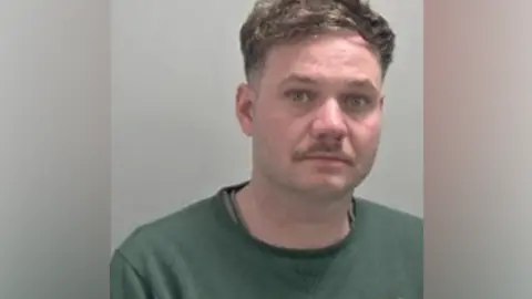A police mugshot of a man with short brown hair, a moustache and a green jumper.