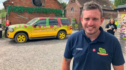 A man standing in a gravel courtyard in front of a brightly coloured dinosaur-themed vehicle and brick buildings.