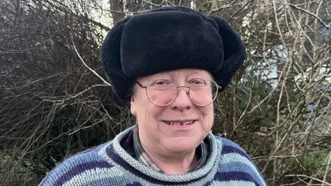 Helen smiles at the camera. She wears glasses, a striped knitted jumper and a furry hat