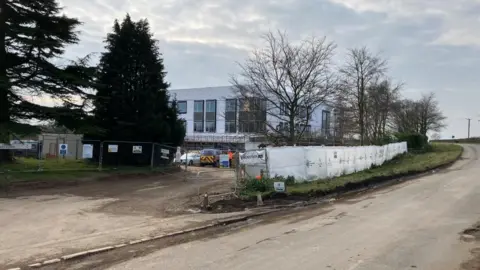 Sam Read/BBC Building of a school, showing a road, trees, and bushes, then a white box building being constructed, with fencing round it, and cars parked outside it. 