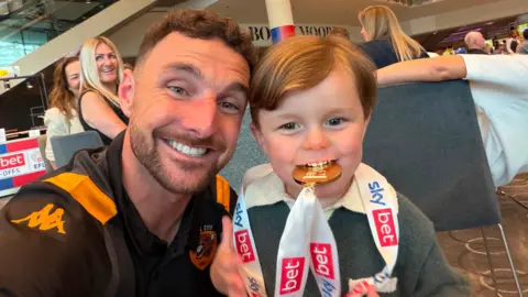 Lewie Coyle A selfie of a man of 30 wearing a Hull City kit and smiling while posing next to his young son who is biting a gold medal while wearing it around his neck.