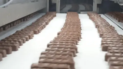 Rows of chocolate bars on a white conveyor belt, under white strip lighting