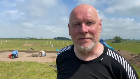 Steve Lowe is a bald man with a grey trimmed beard in his 50s wearing a black top. He's standing in a field and behind him people are digging as part of an archaeological dig 