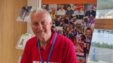 Harrison Galliven / BBC A man wearing a pink shirt sat in front of a poster with faces of people on it.